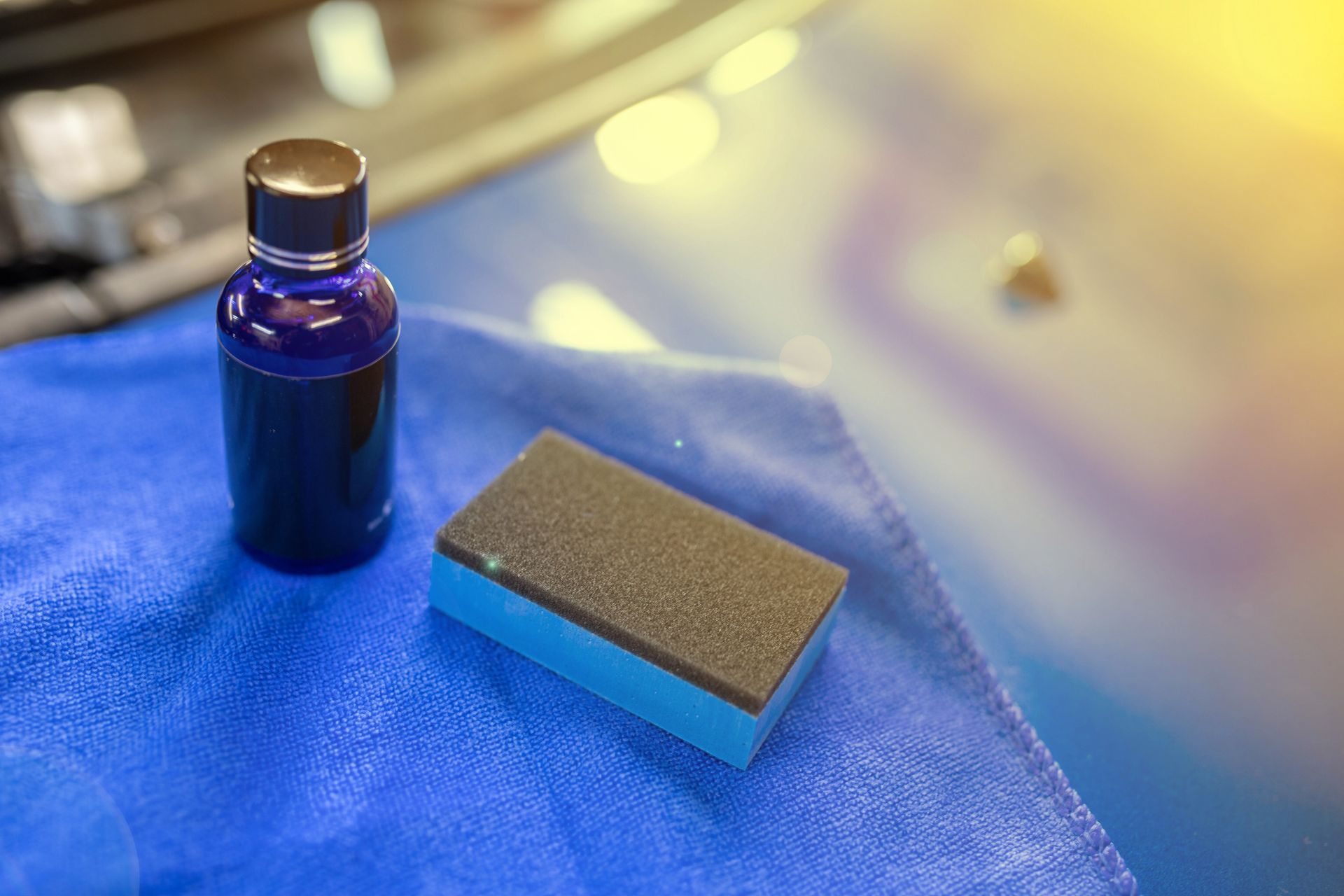 Bottle and applicator sponge on blue microfiber cloth, possibly for car detailing.