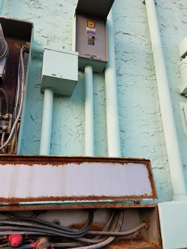 Electrical boxes and conduits mounted on a pale green exterior wall above a rusted metal ledge.