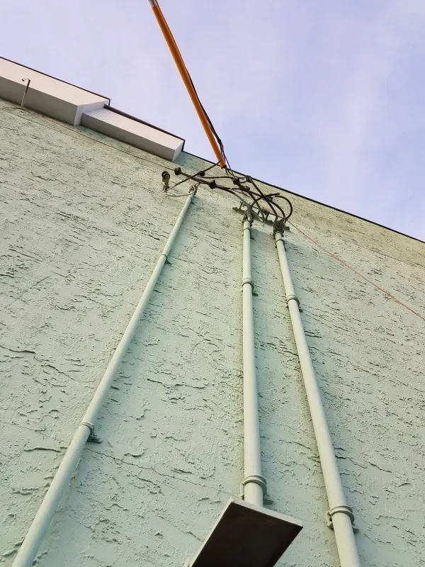 White utility pipes and cables running up a pale green building wall under a cloudy sky.