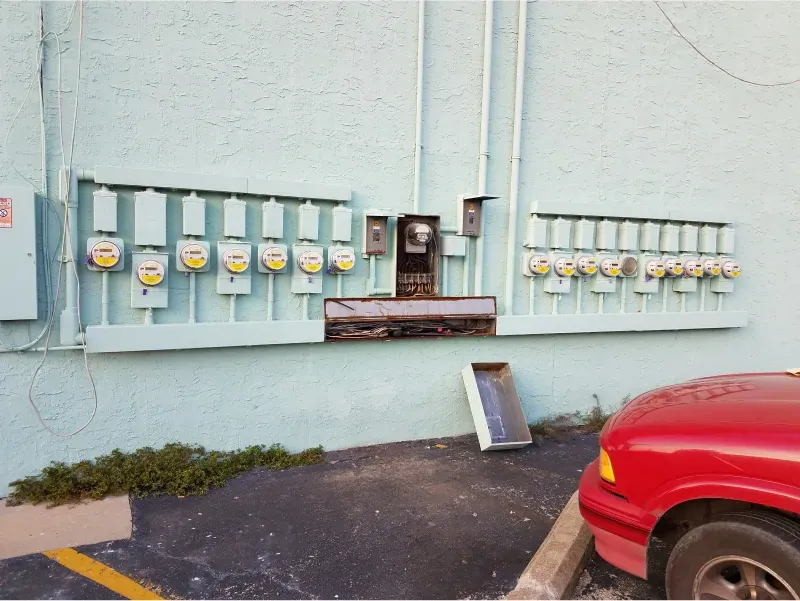 Utility meters and electrical boxes on a pale blue wall beside a red car in a parking area