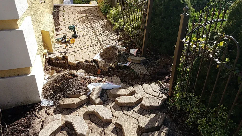 Broken pavers and dirt scattered along a narrow side path beside a house and fence