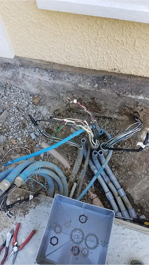 Exposed underground utility cables and conduits in a dirt trench beside a house wall