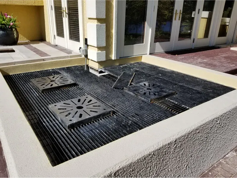 Outdoor drainage pit with black metal grates beside a building entrance
