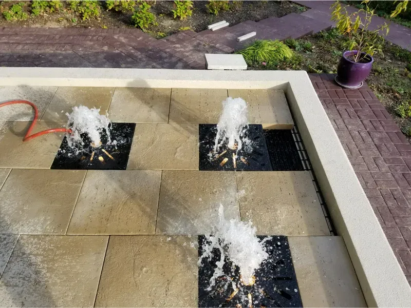 Three small fountains splash in a tiled courtyard with tan stone and a potted plant nearby.