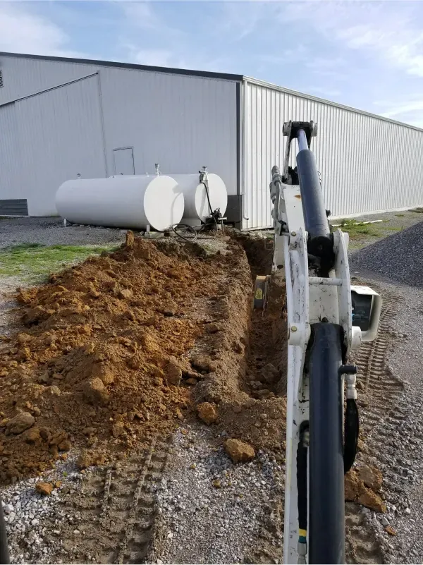 Excavator trench beside white industrial building with dirt pile and propane tanks