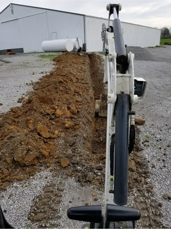 Excavator arm beside a long dirt trench along a gravel lot next to a white industrial building