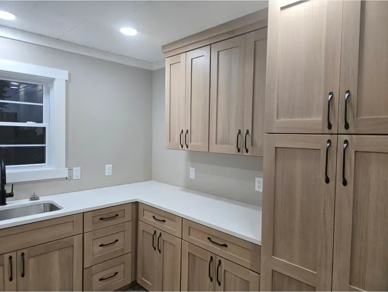 Bright kitchen corner with light wood cabinets, white countertops, and a window above the sink.