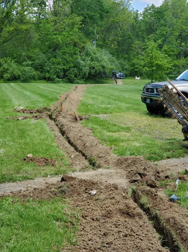 Deep muddy trench cuts across a grassy yard beside a parked green truck and trees