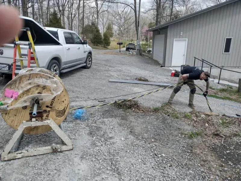Worker using a long tool near a gravel driveway beside a gray building and parked van