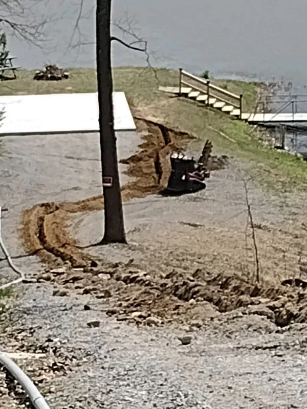 Damaged lakeside path with deep erosion trench, utility pole, and fence along the water’s edge