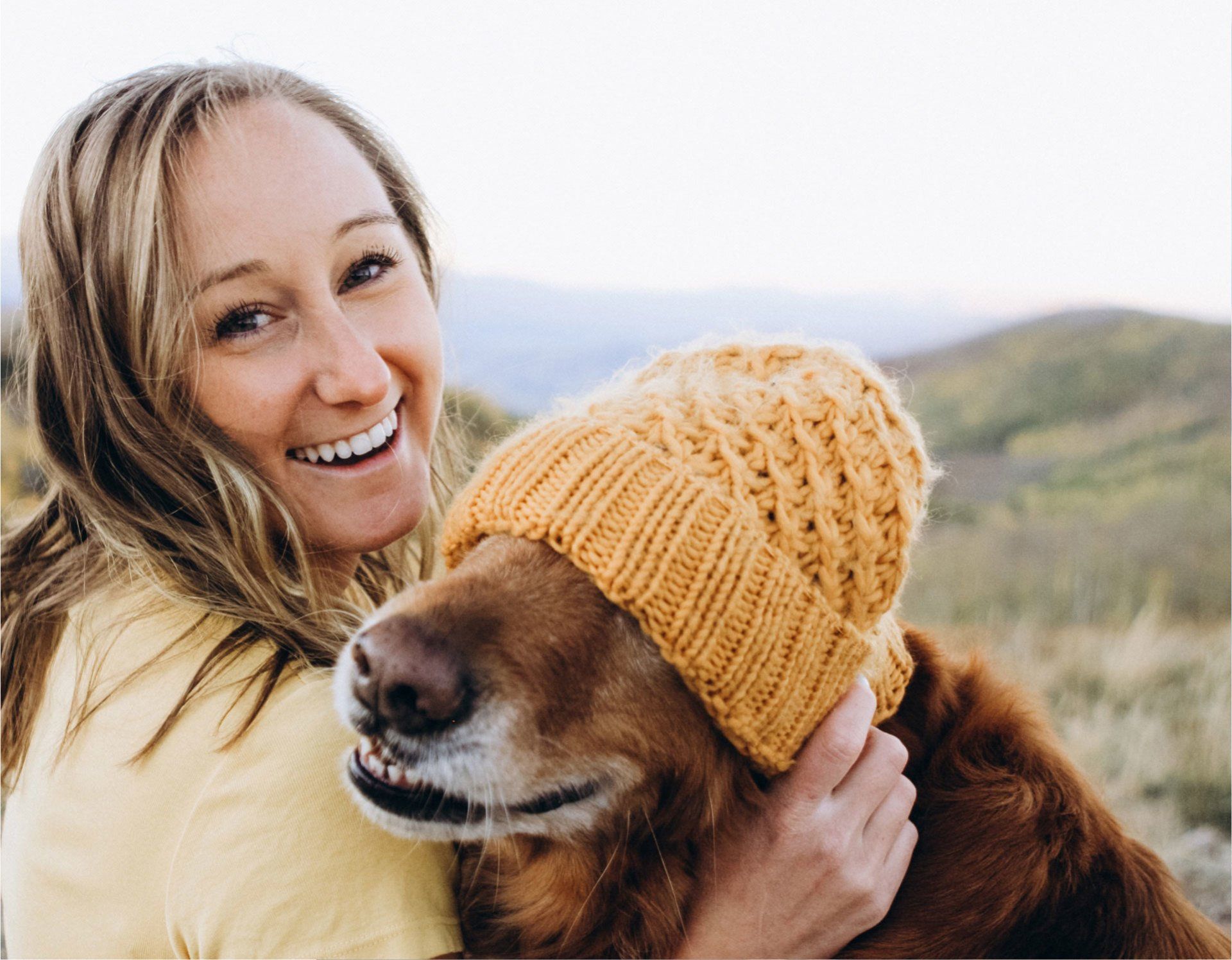 Une femme serrant dans ses bras un golden retriever portant un bonnet jaune en tricot, souriant à l'extérieur.