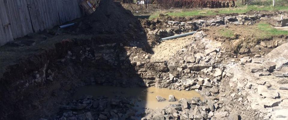 Zone de fouilles avec roches apparentes, terre et eau stagnante, près d'un mur en béton.