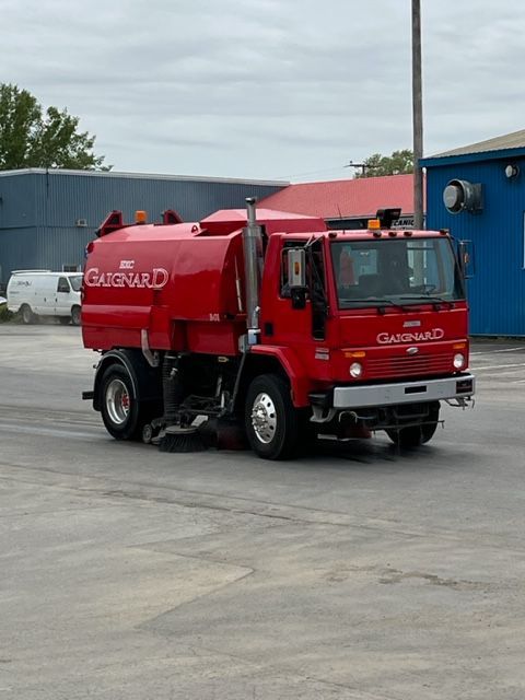 A bright red street sweeper truck moves across a paved parking lot near a blue building.