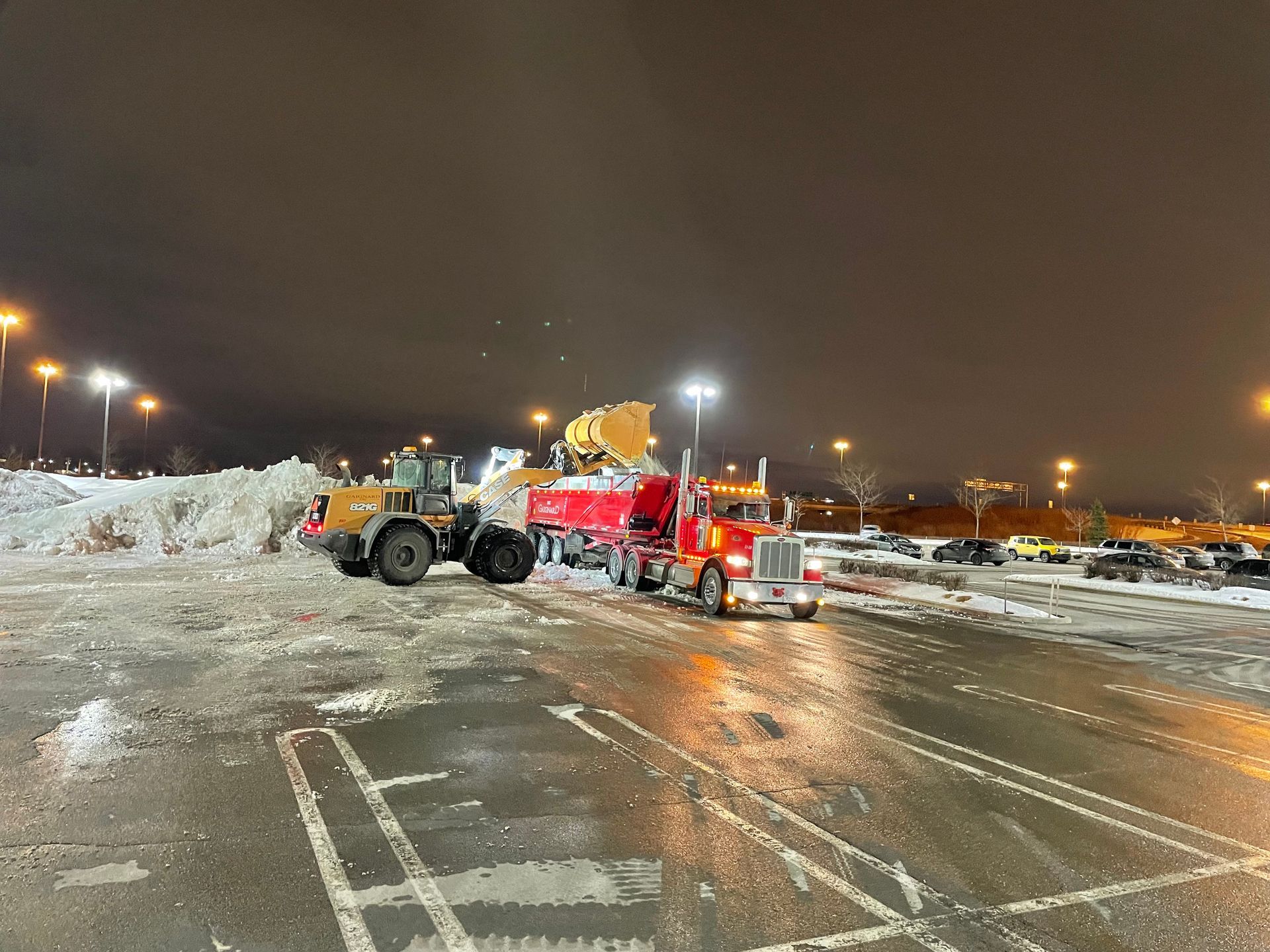 Déneigement de nuit : une chargeuse remplit un camion-benne rouge de neige sur un parking éclairé par des lampadaires.