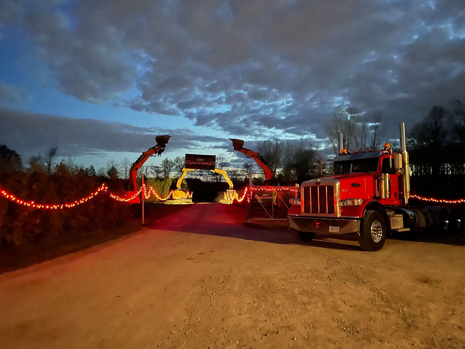 Un semi-remorque rouge est stationné près d'une arche illuminée aux couleurs de l'événement, au crépuscule, sous un ciel nuageux.