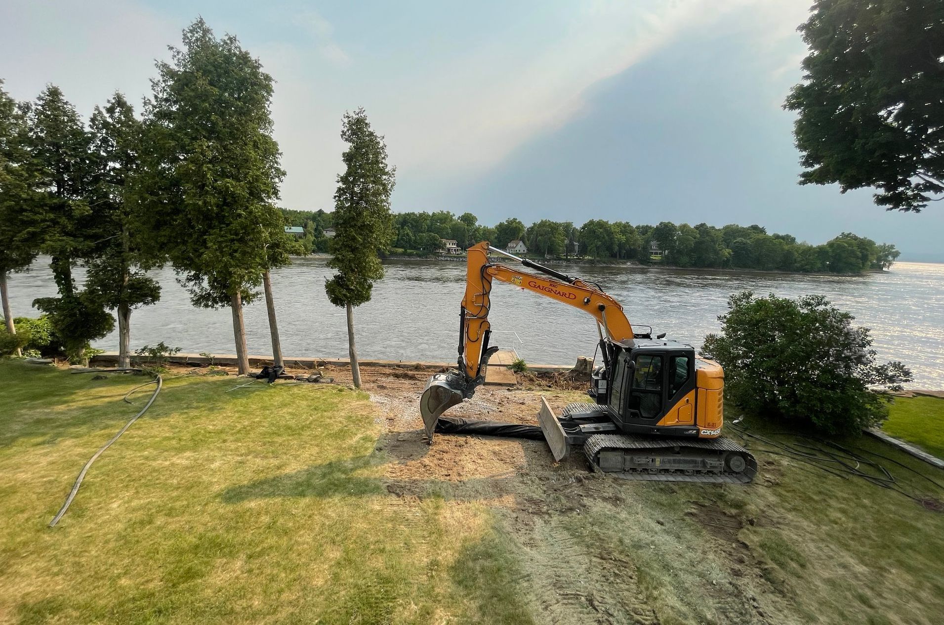 Pelleteuse sur une berge herbeuse au bord de l'eau, arbres en arrière-plan, journée ensoleillée.
