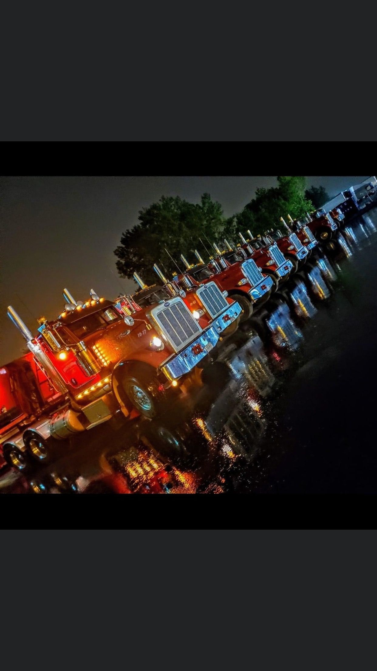 Une rangée de semi-remorques illuminées stationnées la nuit ; les lumières rouges et bleues se reflètent sur le sol humide.