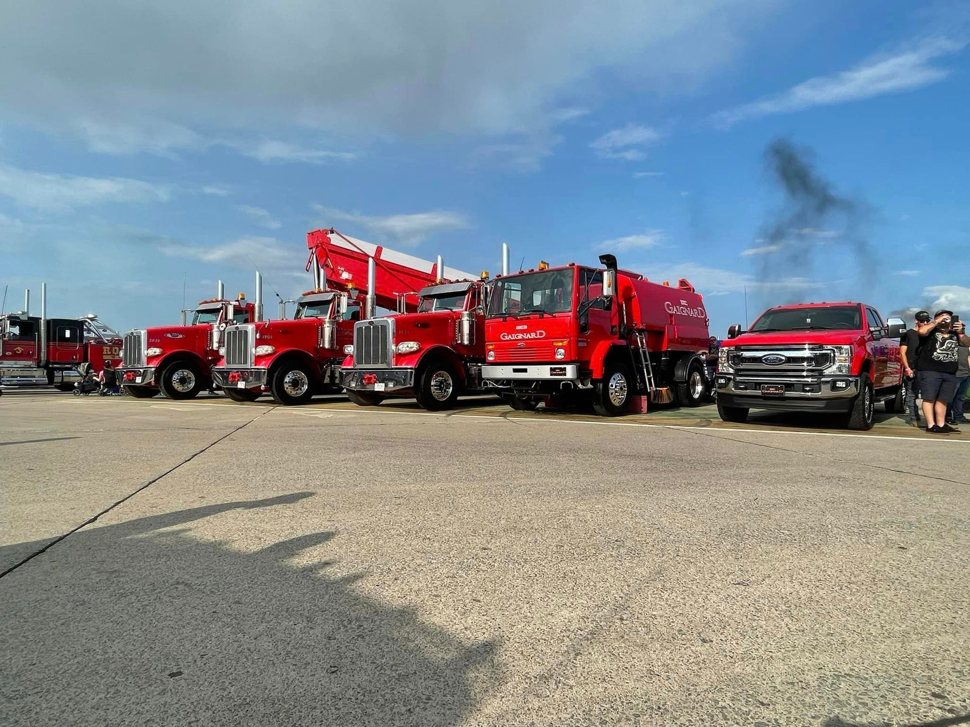 Des camions de pompiers rouges alignés sur une surface en béton sous un ciel bleu, avec quelques personnes en arrière-plan.