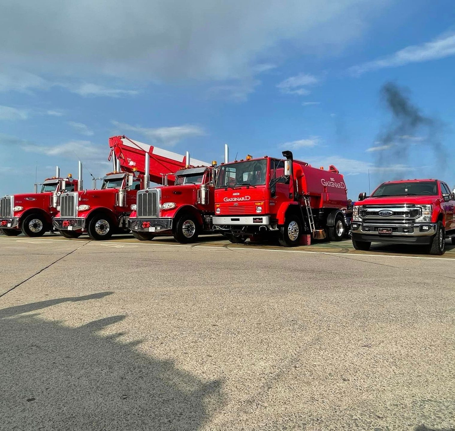 A row of red service trucks parked on an asphalt lot under a blue sky with faint smoke in the distance.