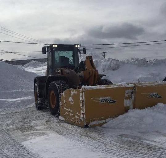 A yellow front-end loader with a wide snow pusher attachment clearing a large snowy lot under a cloudy sky.