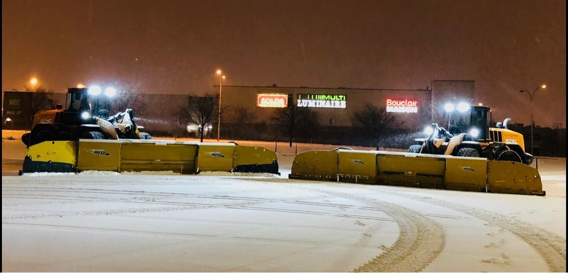 Deux chasse-neige dégagent une zone enneigée de nuit. Leurs lames jaunes sont abaissées. Leurs phares puissants éclairent la scène.