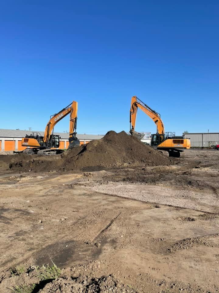 Two yellow excavators face each other across a large pile of dirt on a sunny construction site.