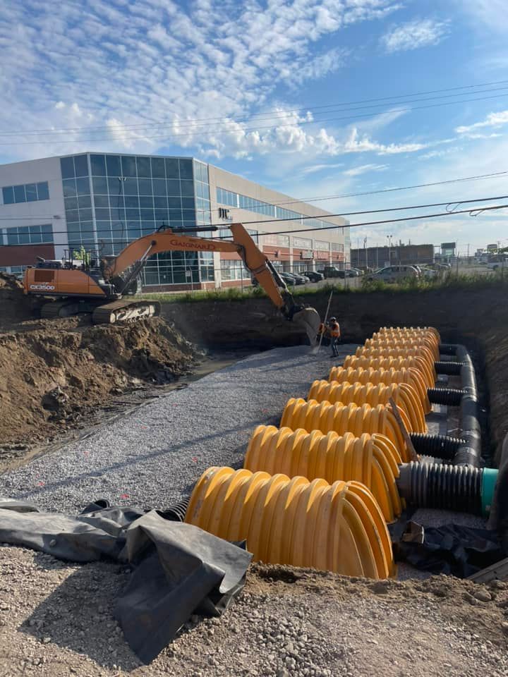 Chantier de construction avec une excavatrice, des structures de drainage en plastique jaune enterrées et un bâtiment en arrière-plan.