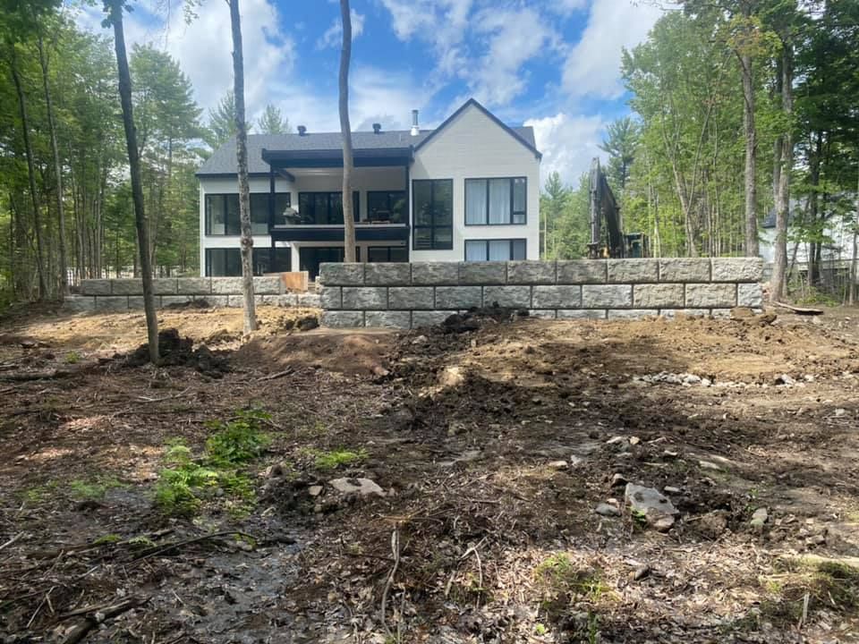 A modern white house with large windows sits behind a tiered gray stone retaining wall in a partially cleared wooded lot.