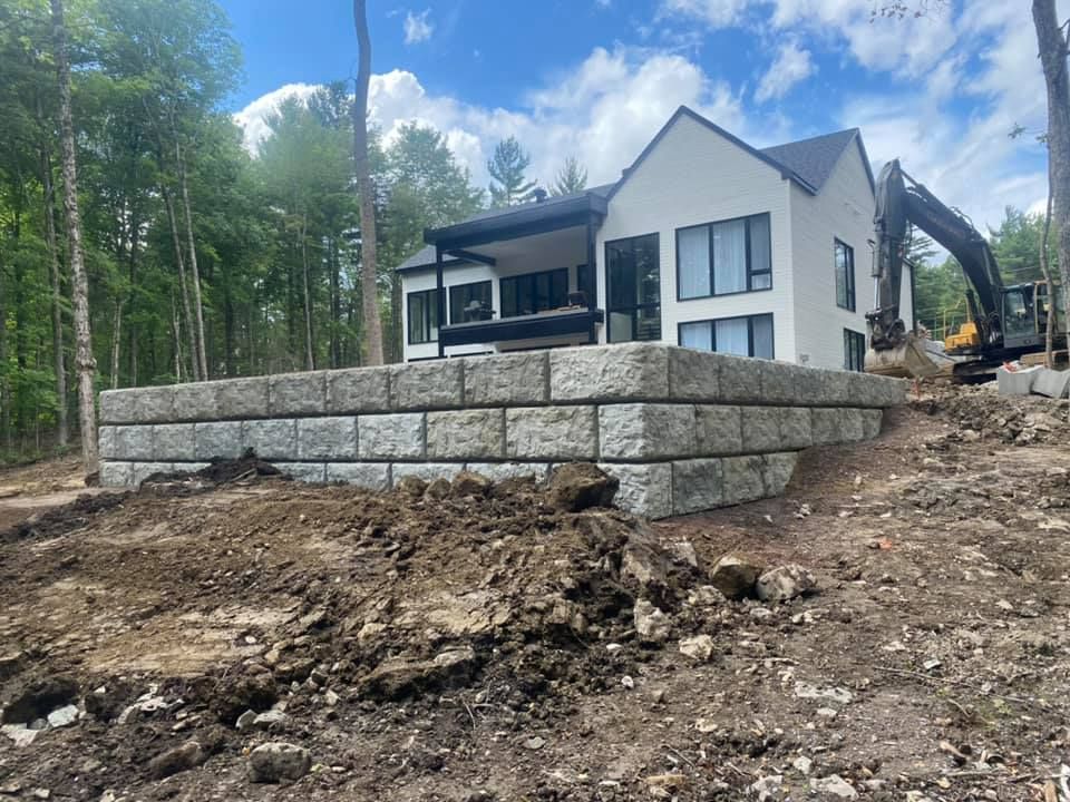 Une maison blanche moderne aux grandes fenêtres surplombe un mur de soutènement en pierre grise. Une pelleteuse se trouve à proximité.