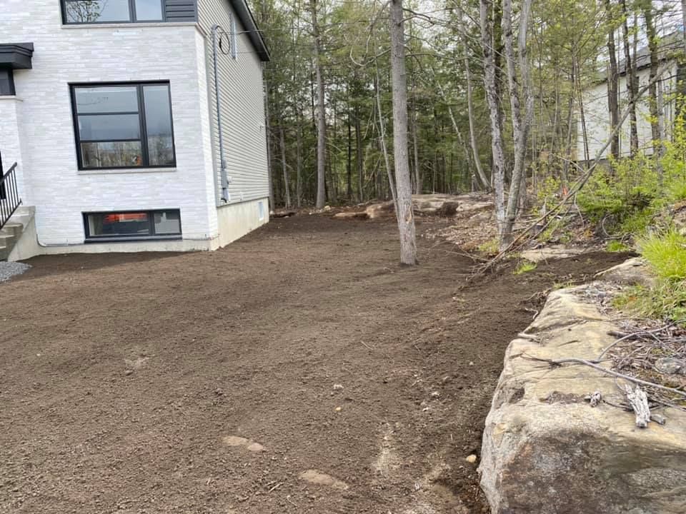 Une maison avec un jardin récemment retourné, jouxtant une zone boisée parsemée de gros rochers.