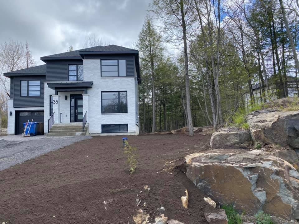Maison moderne à deux étages avec des boiseries noires et une façade en briques blanches, située sur un terrain légèrement en pente avec une formation rocheuse et des arbres.