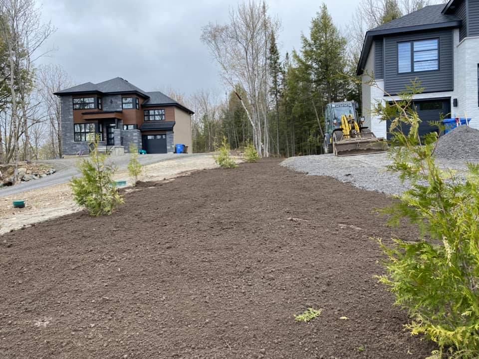 De la terre fraîchement excavée devant deux maisons modernes, une petite pelleteuse et des arbres ; journée nuageuse.