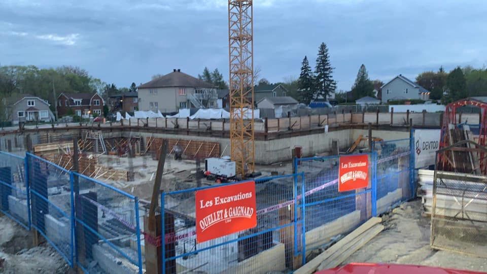 Chantier de construction avec grue, fosse clôturée et panneaux « Les Essentielles », maisons en arrière-plan.