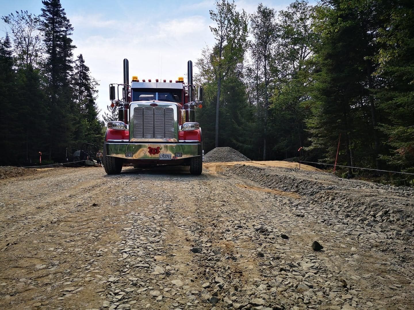Un semi-remorque sur un chemin de gravier s'approche de la caméra, avec des arbres en arrière-plan.