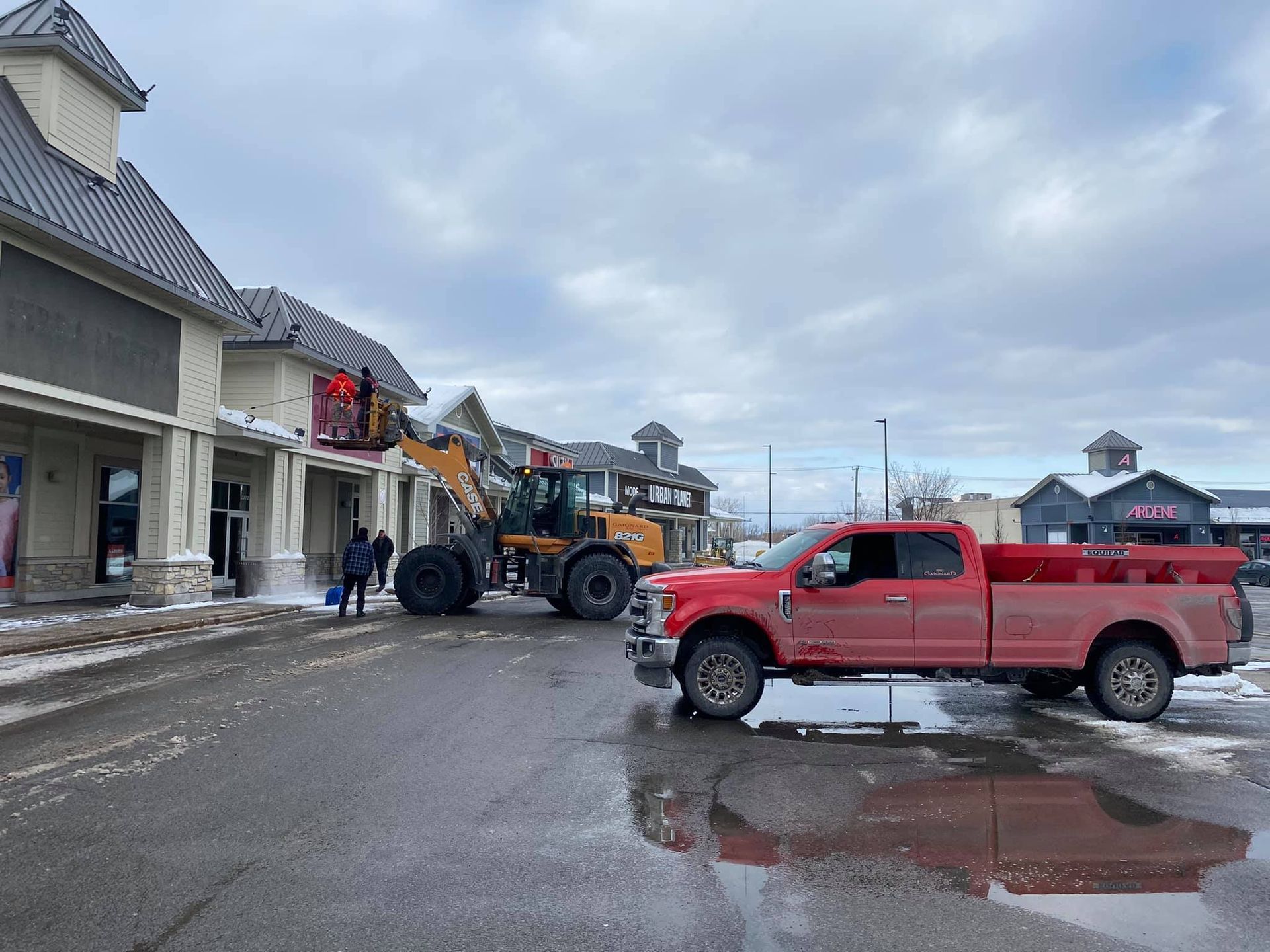 Un camion rouge est stationné près d'un bâtiment tandis qu'une chargeuse frontale déneige le toit. Ciel nuageux.