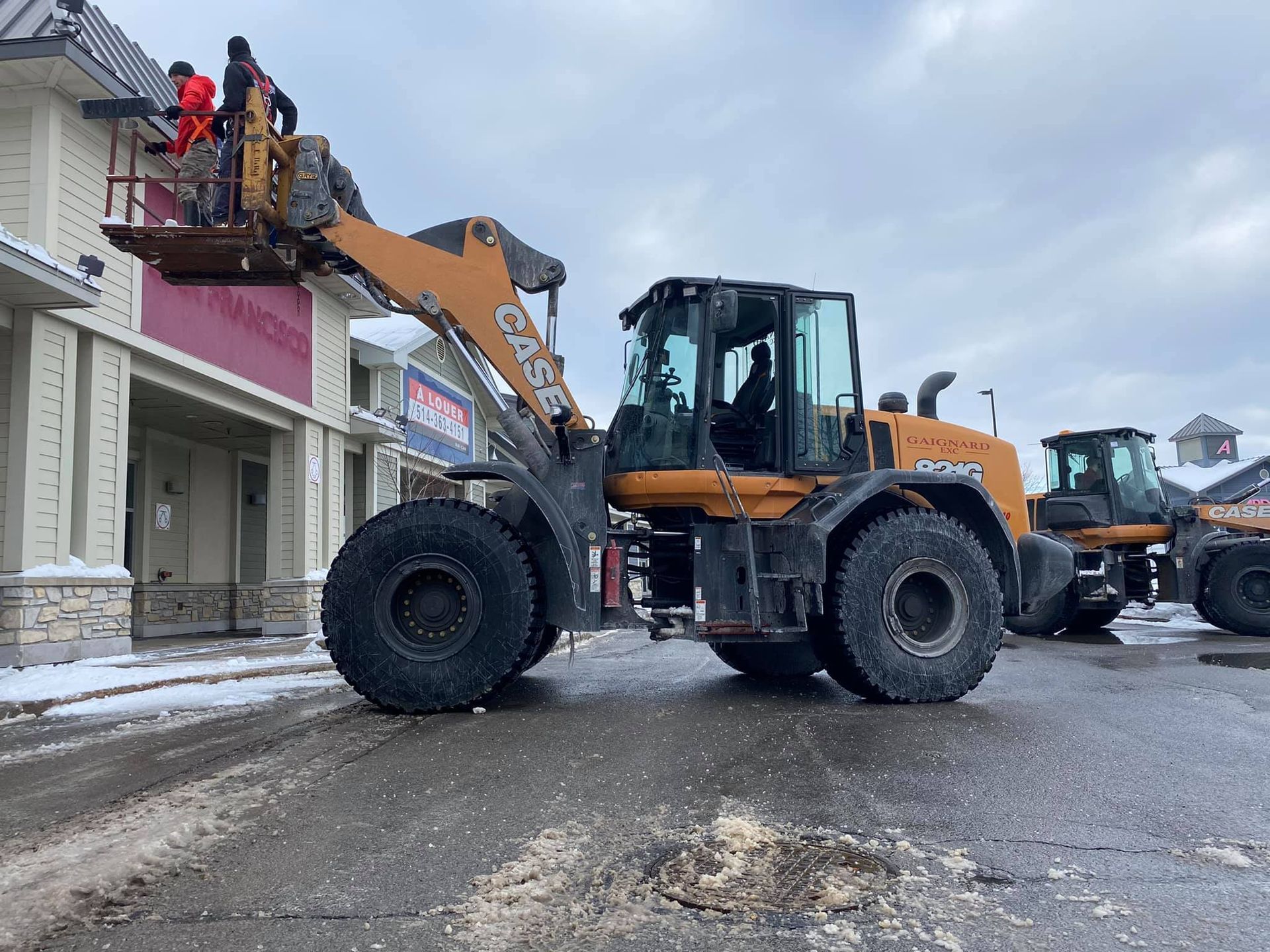 Workers in a construction basket attached to an orange Case loader, parked outside a building on a winter day.