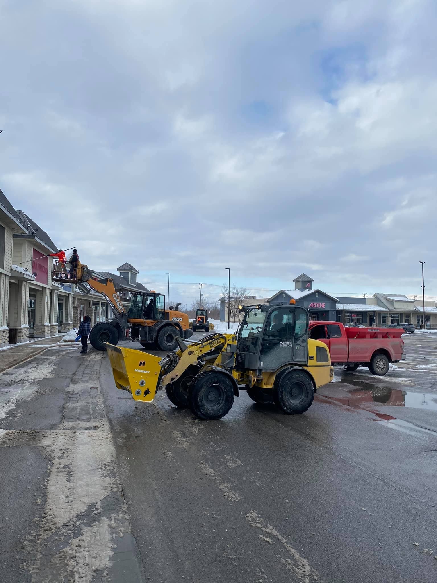 Une chargeuse jaune déneige une rue d'une ville ; d'autres engins et bâtiments sont visibles. Ciel couvert.