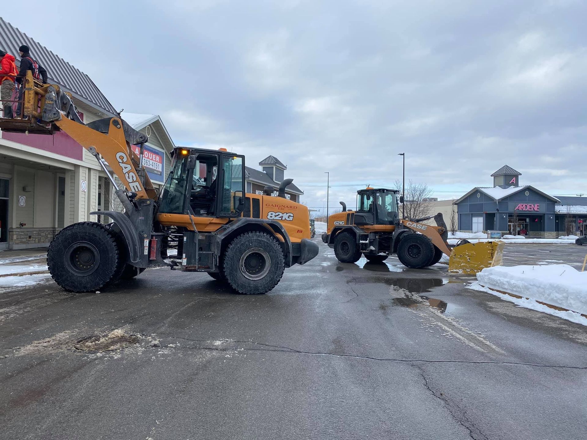 Deux chargeuses sur pneus Case déneigent un bâtiment commercial. L'une est équipée d'un godet surélevé, l'autre d'une lame à neige.