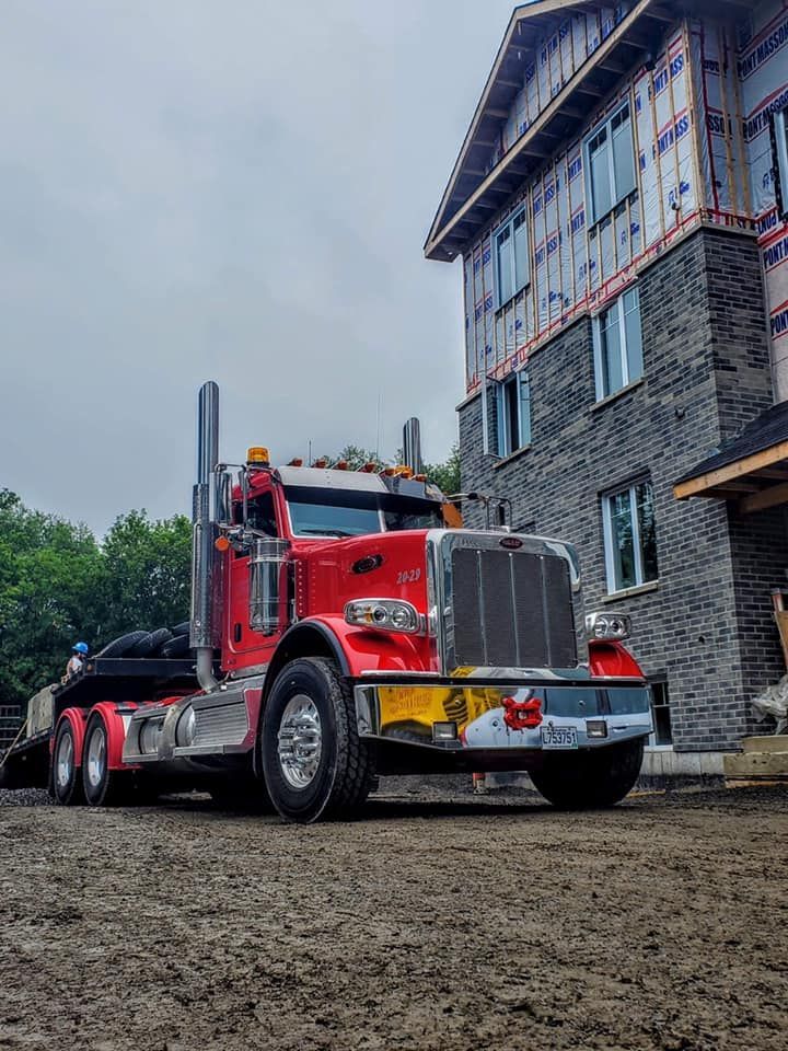 Un semi-remorque rouge est stationné sur un chemin de terre devant un bâtiment en construction.