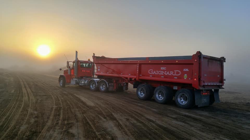 Un semi-remorque rouge avec une benne basculante roule sur une route sablonneuse, soleil et brouillard en arrière-plan.