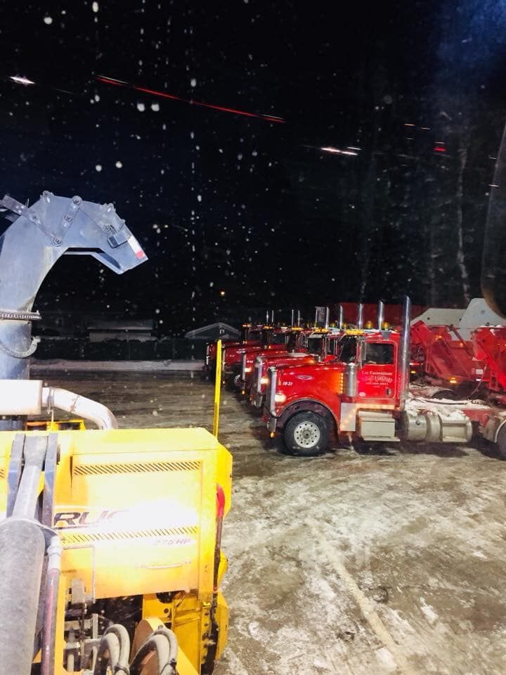 Une file de semi-remorques rouges stationnées de nuit, sous la neige. Une souffleuse à neige jaune est visible au premier plan.