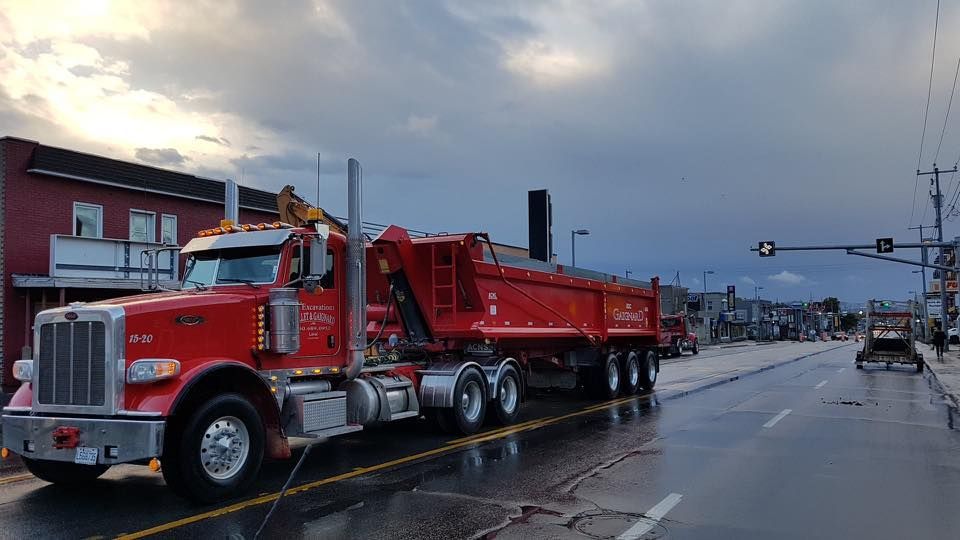 Camion-benne rouge sur une rue mouillée. Ciel sombre au-dessus ; bâtiments et autres véhicules visibles.