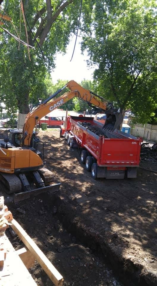 Une excavatrice charge un camion-benne rouge de terre sombre dans un décor extérieur arboré.