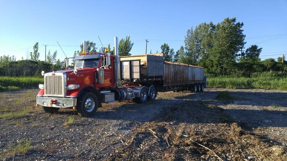 Un semi-remorque rouge tirant une longue remorque rouillée est stationné sur du gravier, sous un ciel bleu, près d'arbres verts.