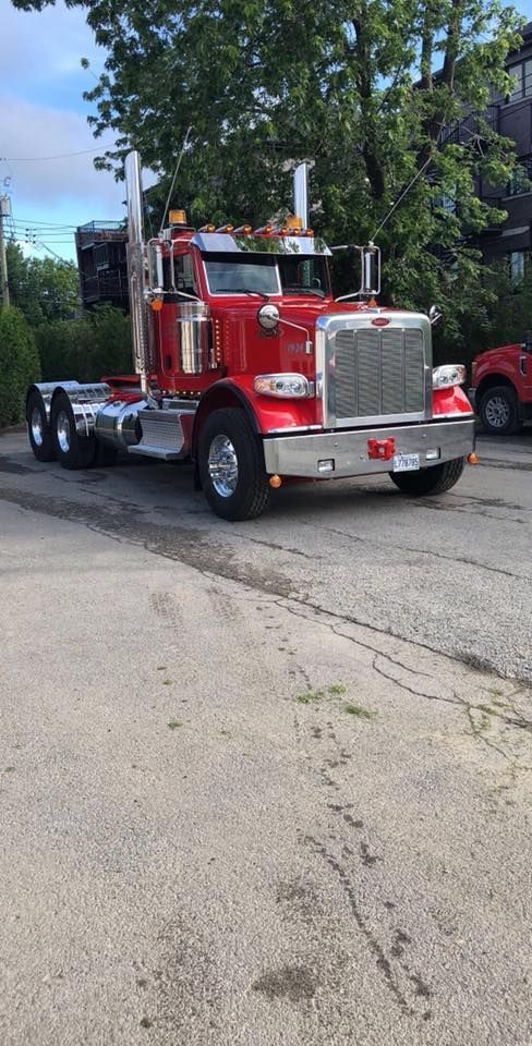 Un semi-remorque Peterbilt rouge est stationné sur une chaussée fissurée ; des arbres et un bâtiment se trouvent en arrière-plan.
