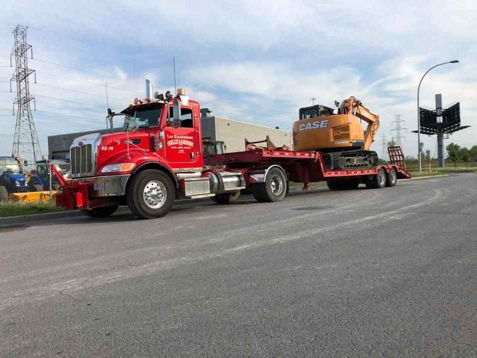 Un semi-remorque rouge transportant une pelle mécanique Case sur une remorque à plateau est stationné sur l'asphalte.