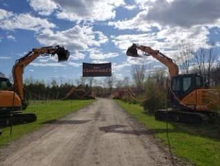 Deux pelleteuses orange encadrent un chemin de terre surmonté d'une banderole, sous un ciel nuageux.