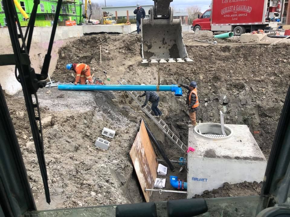 Chantier avec ouvriers, excavatrice et grand trou. Des ouvriers manipulent un tuyau bleu près d'une structure en béton.