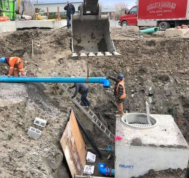 Construction workers install blue utility pipes at a site with an excavator, a red service truck, and a concrete basin.