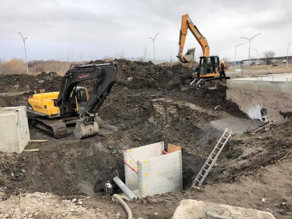 Chantier de construction avec deux excavatrices creusant une fosse boueuse ; une échelle est appuyée contre une structure en béton.
